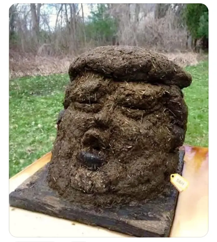 A crudely sculpted bust resembling Donald Trump, made entirely of dirt and straw, sits on a wooden base outdoors with a…