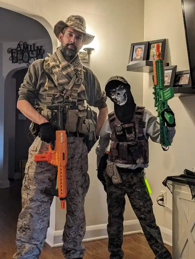 A man and a young boy are posing indoors in full tactical gear, including camouflage pants, vests, and head coverings. The…