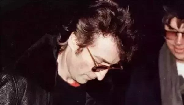 A grainy photo shows a man with dark hair and sunglasses, wearing a black leather jacket and looking down, while another man…