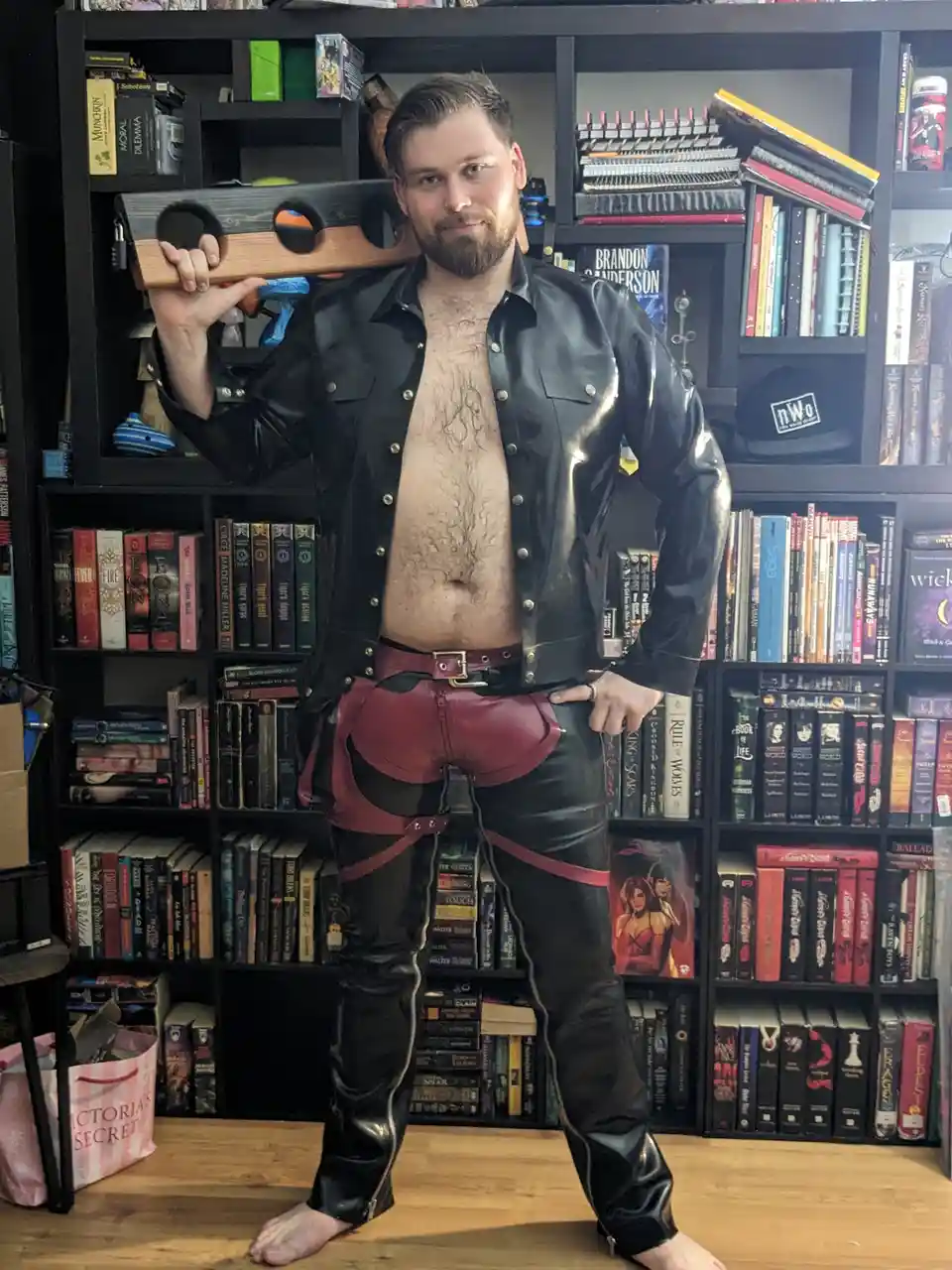 A man with a beard and bare chest stands in front of a bookshelf, wearing a shiny black leather jacket, black leather pants…