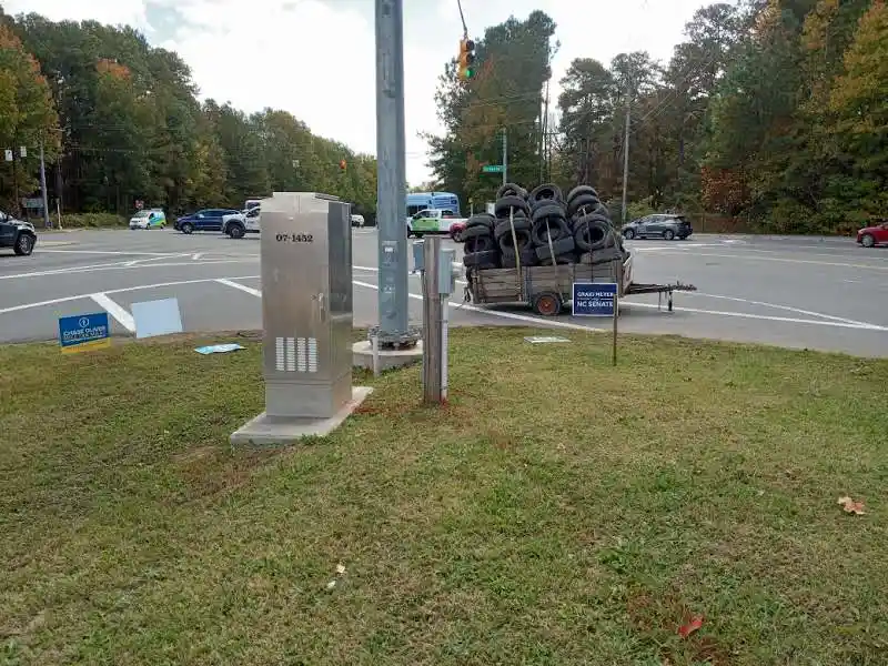 A roadside scene shows a utility box and a trailer piled high with used tires, both situated near a traffic intersection. A…