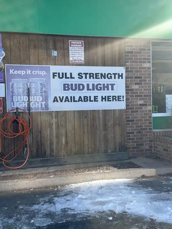 A large white banner on a wooden wall of a store advertises 'FULL STRENGTH BUD LIGHT AVAILABLE HERE!' in bold black and…