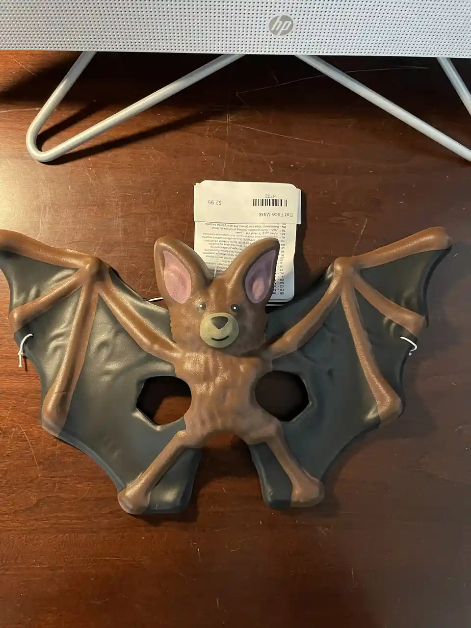 A brown plastic bat-shaped face mask with black wings and pink inner ears rests on a wooden desk, with a price tag reading…