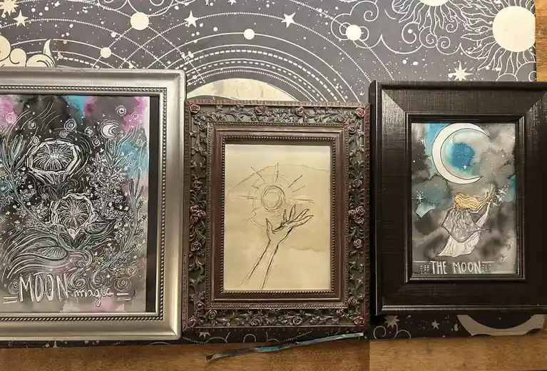 Three framed artworks sit on a wooden surface against a celestial-patterned backdrop. The left frame shows a cosmic design…