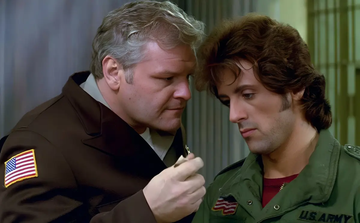 two men in military-style jackets stand close together, one in a brown coat with an american flag patch on the sleeve, the…
