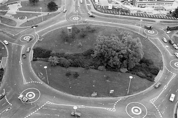 A black-and-white aerial photo of a large, circular road junction with multiple lanes and cars. The central island has trees…