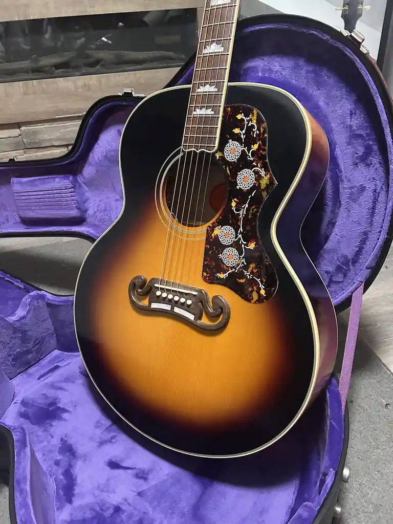 An acoustic guitar with a sunburst finish and ornate floral inlay sits inside a purple velvet case. The guitar's bridge is…