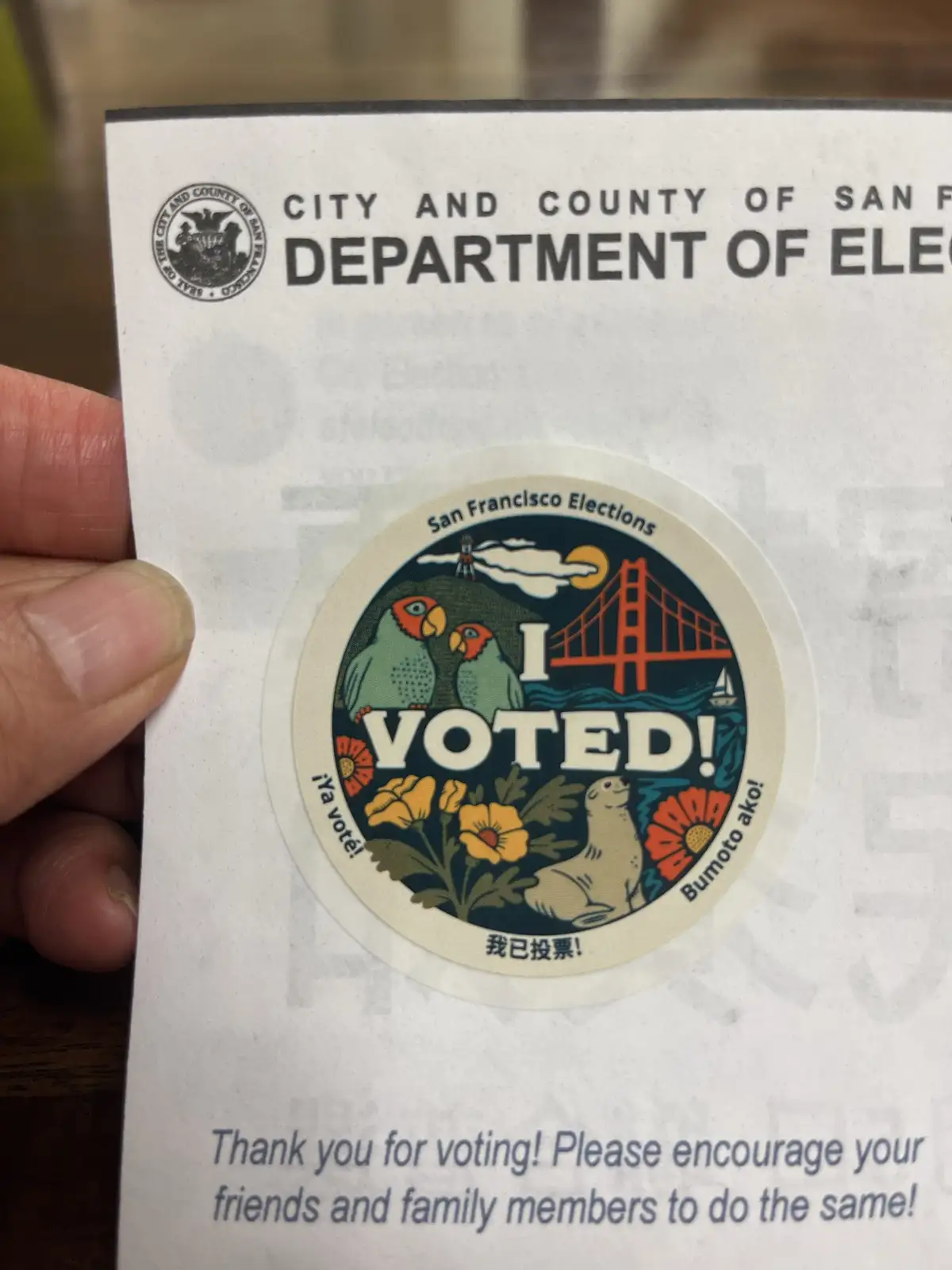 A hand holds a San Francisco Department of Elections 'I VOTED!' sticker, which features the Golden Gate Bridge, two parrots…