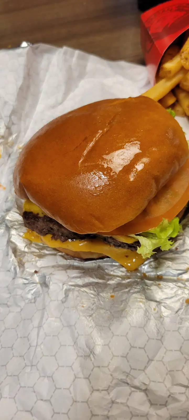 A close-up photo of a cheeseburger with a glossy top bun, melted cheese, tomato, and lettuce, sitting on foil with fries…