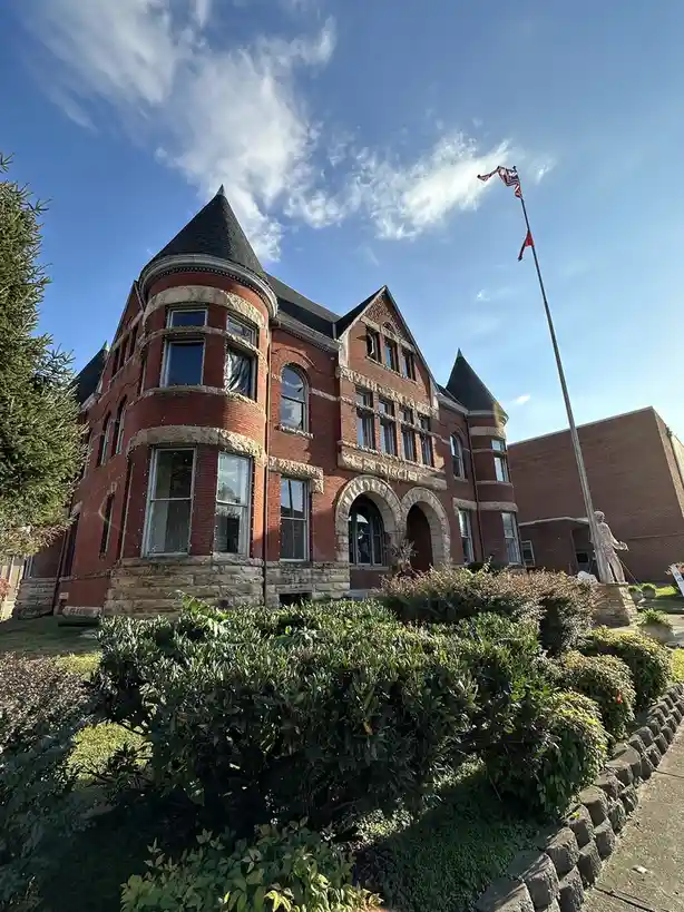 A low-angle photo shows a large, red-brick building with stone trim, turrets, and arched windows under a blue sky with wispy…
