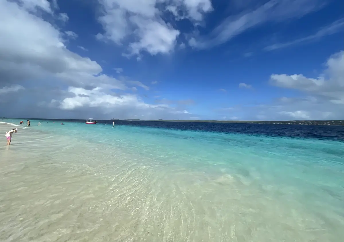 A serene tropical beach scene with clear turquoise water, white sand, and a blue sky with scattered clouds. Several people…