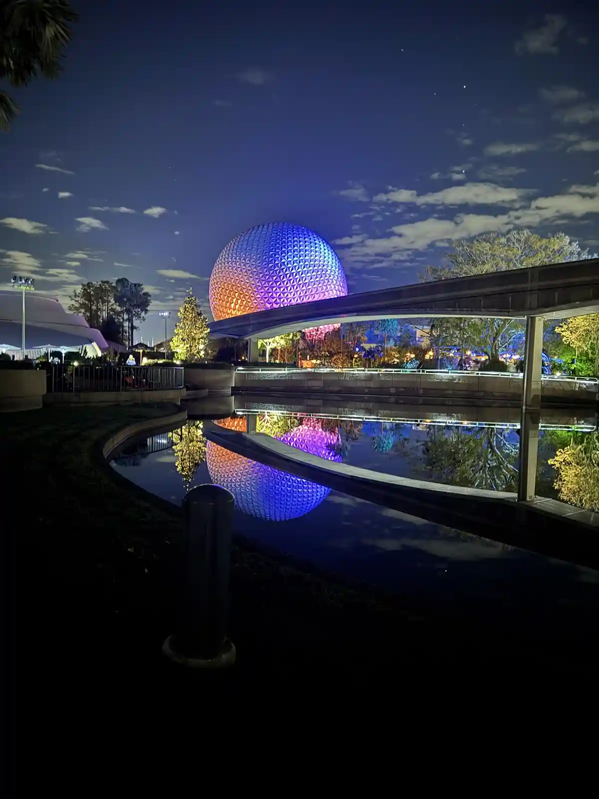 A nighttime photo of the iconic Spaceship Earth geosphere at Epcot, illuminated with blue and orange lights, reflected in a…