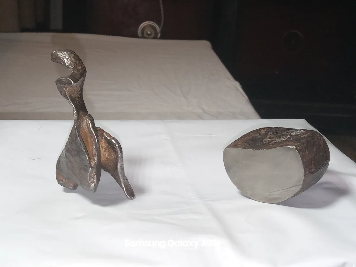 Two rusty, oddly shaped metal objects sit on a white cloth. One resembles a twisted, abstract figure, the other is a…
