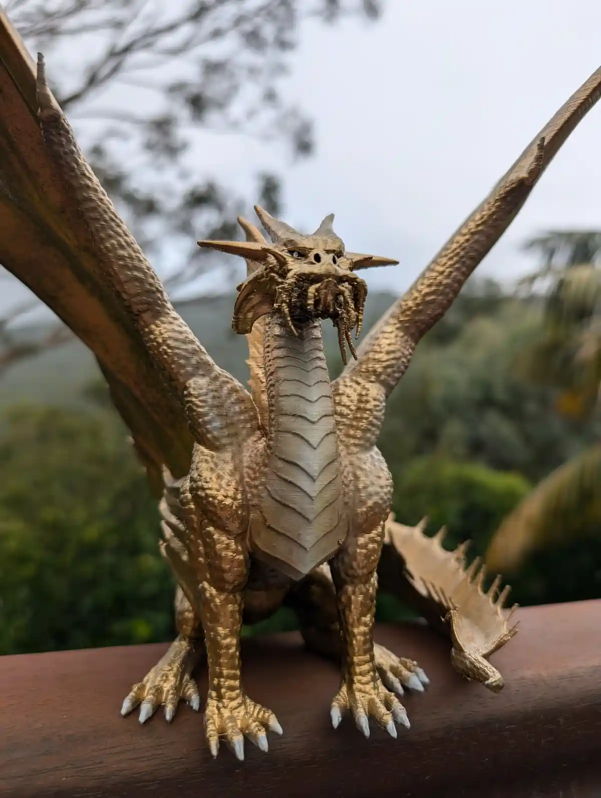 A golden, 3D-printed dragon statue with spread wings and sharp claws stands on a wooden surface outdoors, with trees and sky…