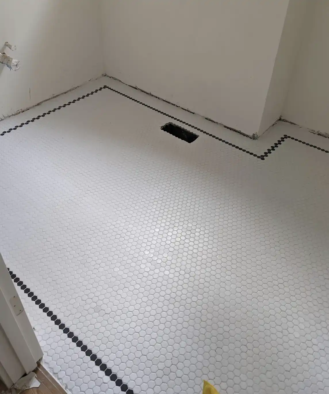 A photo of a bathroom floor covered in white hexagonal tiles with a black border, showing a large rectangular hole in the…