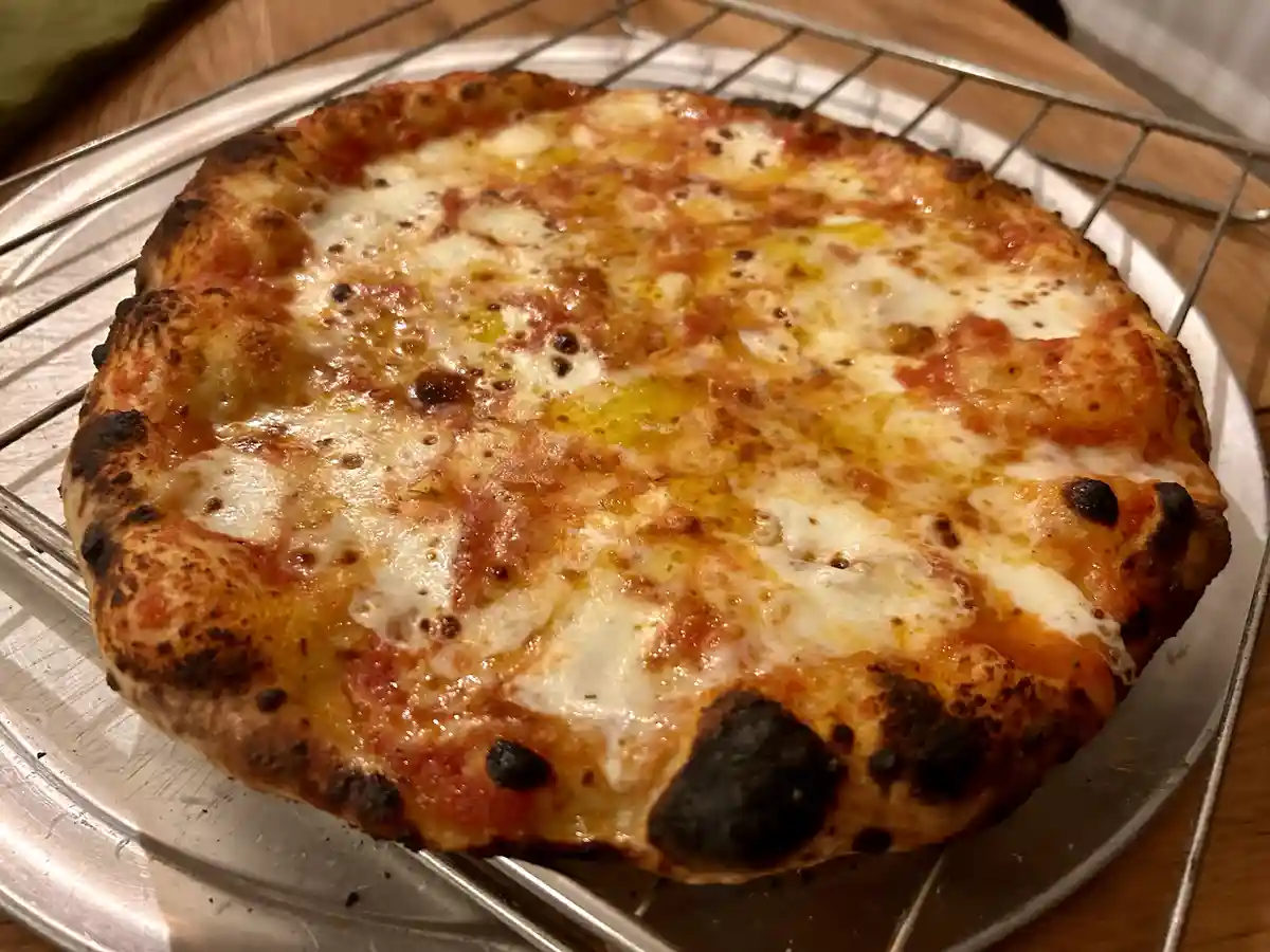 A close-up photo of a freshly baked pizza with a charred crust and melted cheese, resting on a metal cooling rack atop a…