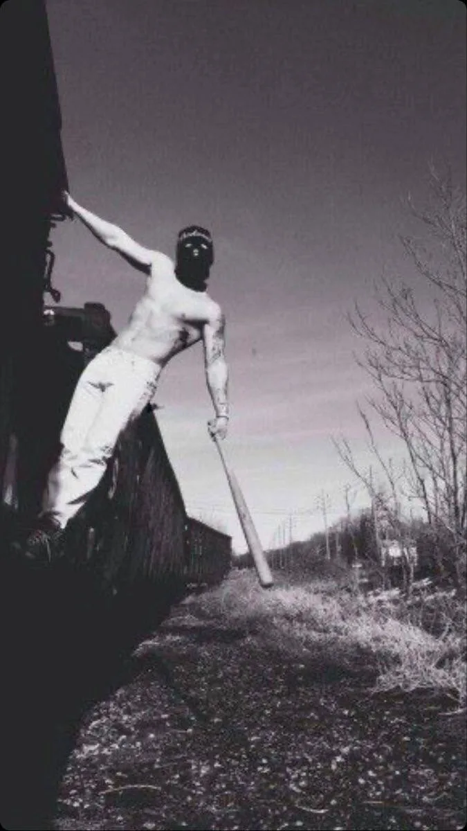 A black-and-white photo shows a shirtless man wearing a black ski mask, standing on the side of a moving train while holding…