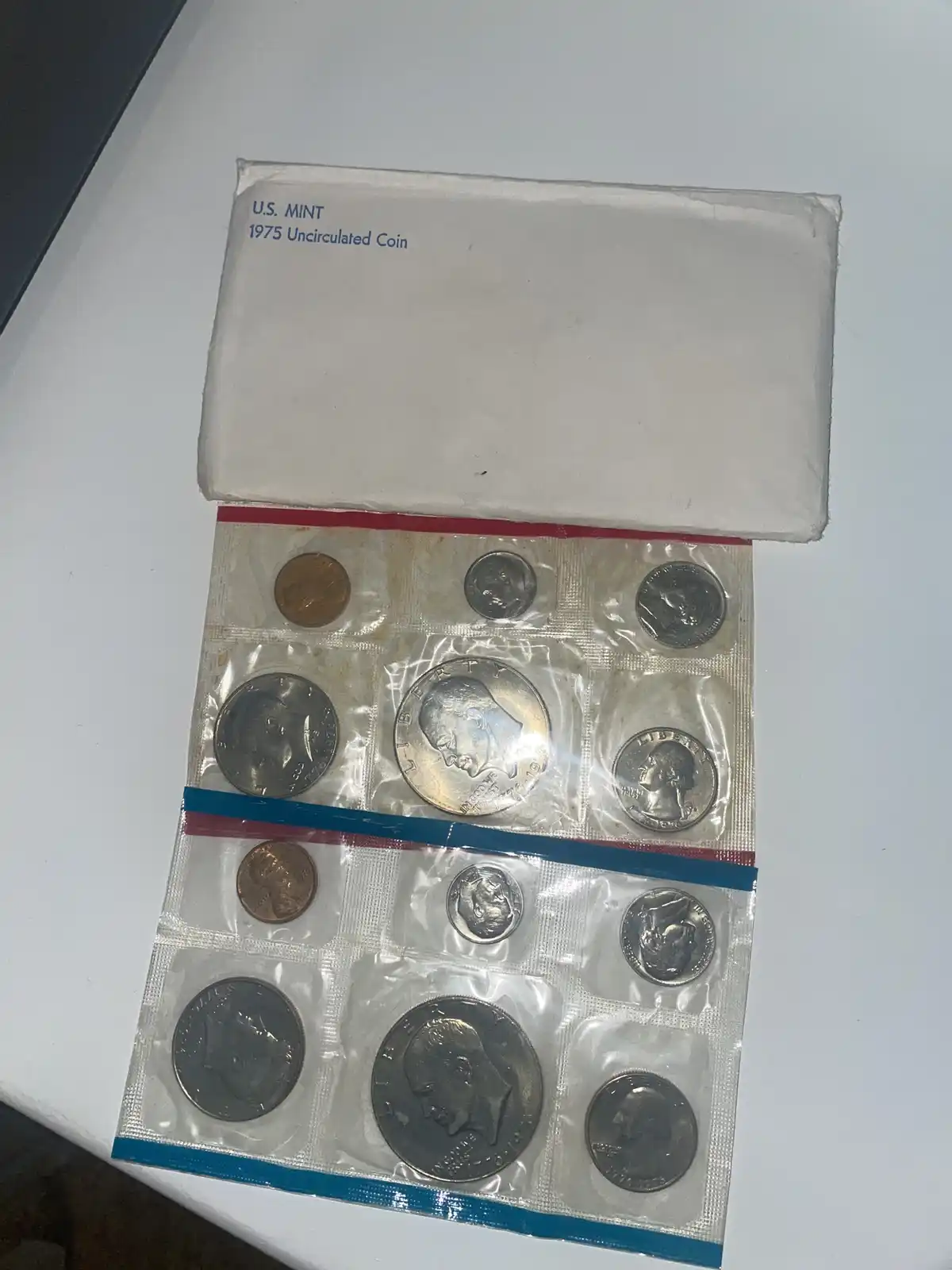 A white cardboard box labeled 'U.S. MINT 1975 Uncirculated Coin' is open, revealing a plastic coin holder with 12 U.S. coins…