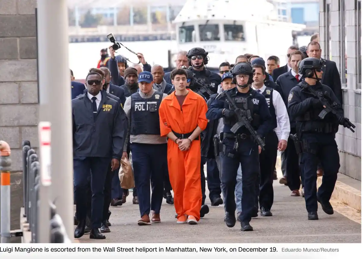 A man in an orange prison jumpsuit is being escorted by numerous armed NYPD officers along a waterfront walkway. The…