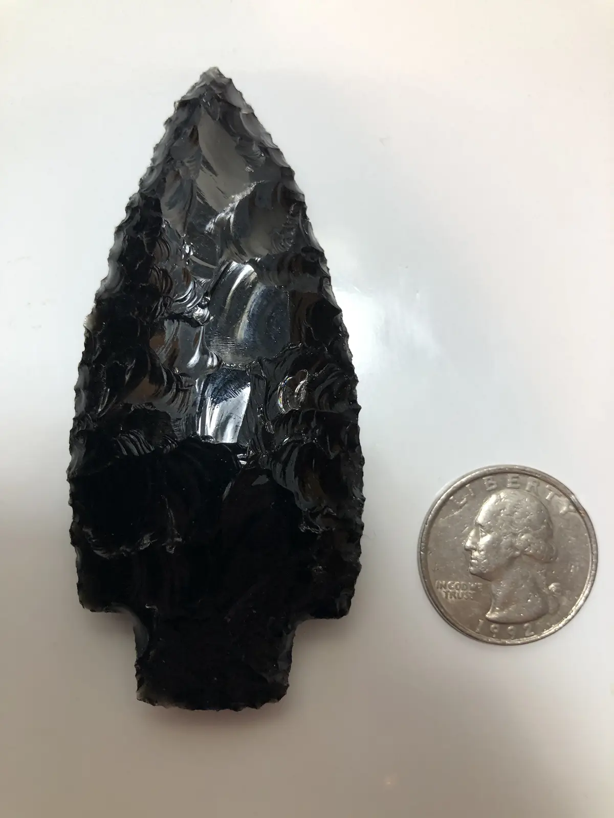 A glossy black obsidian arrowhead with a pointed tip and flaked edges lies next to a 1992 U.S. quarter on a plain white…