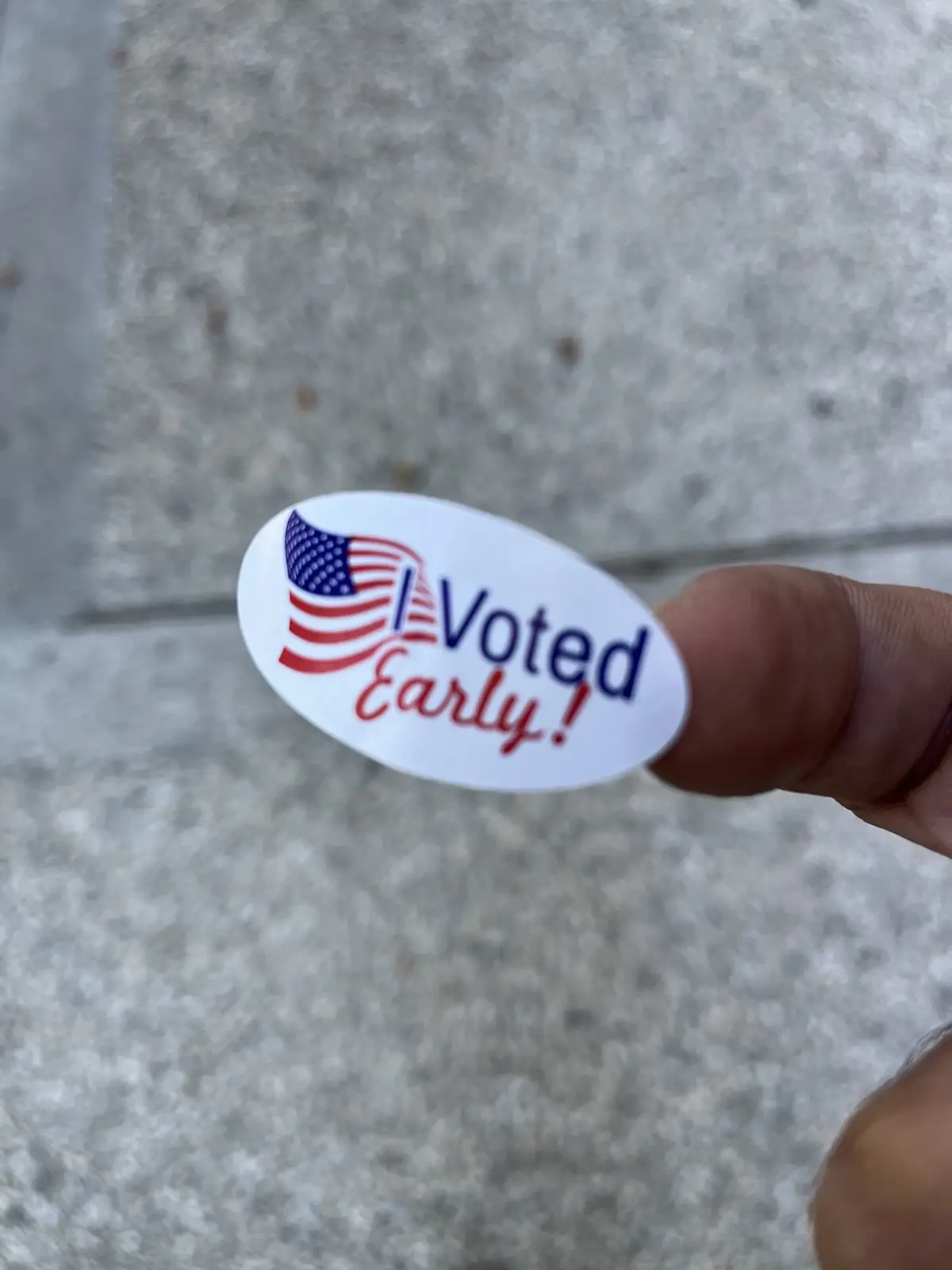 A close-up photo shows a person's thumb holding a white oval sticker with a stylized American flag and the text 'I Voted…
