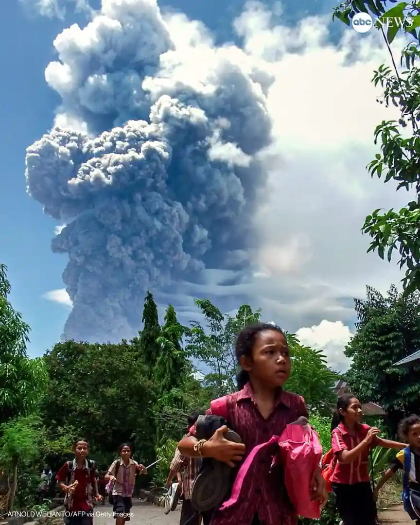 A photo shows children walking outdoors with a massive volcanic eruption and plume of ash dominating the sky behind them…