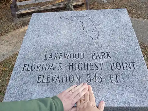 A close-up photo shows a gray granite marker in a park with engraved text reading 'LAKEWOOD PARK', 'FLORIDA'S HIGHEST…