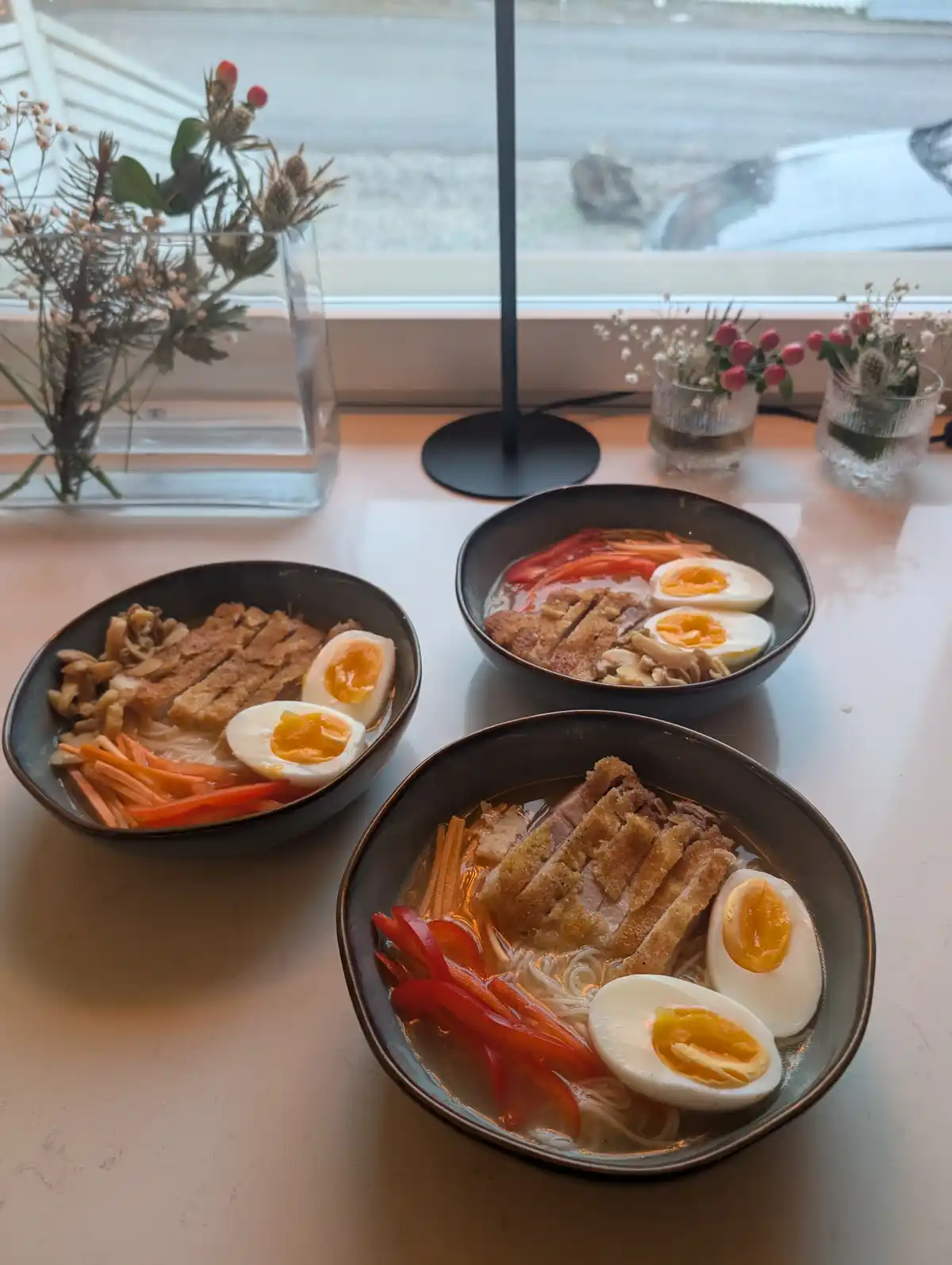 Three bowls of ramen sit on a table near a window, each containing sliced breaded pork, noodles, sliced red peppers…