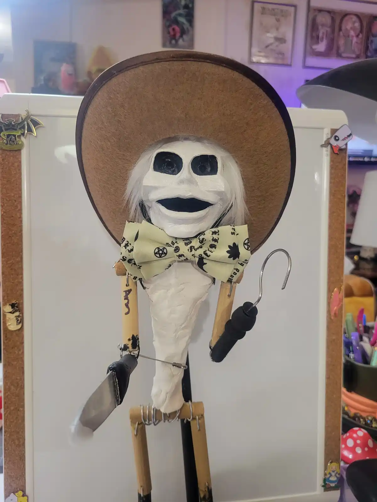 A handmade, white-painted skeleton puppet with a wide-brimmed brown hat and a patterned bow tie hangs from a whiteboard. It…