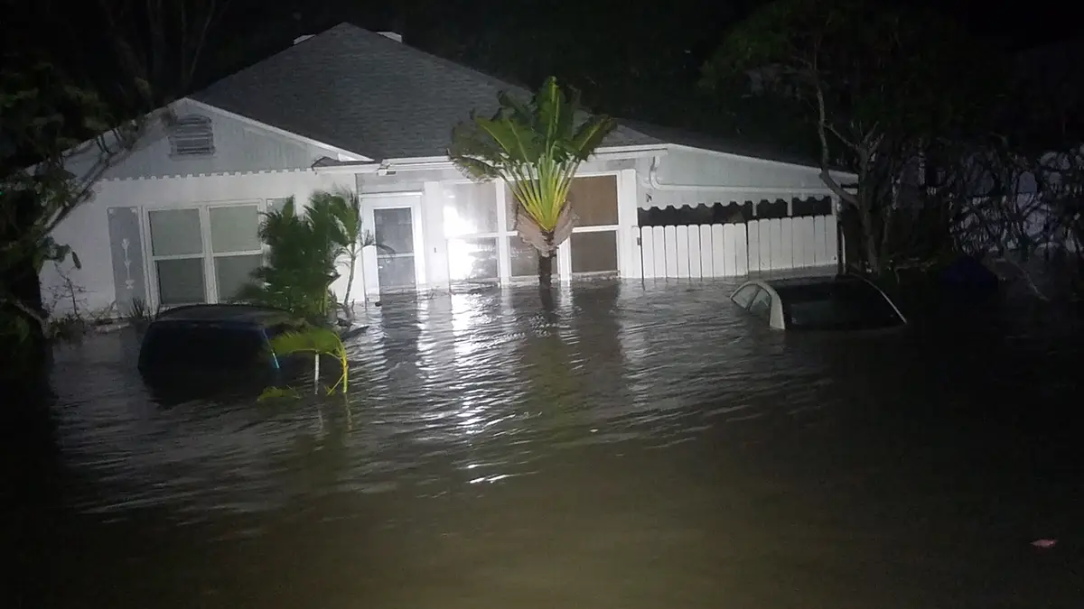 A nighttime photo shows a single-story white house partially submerged in floodwaters, with two cars also underwater in the…