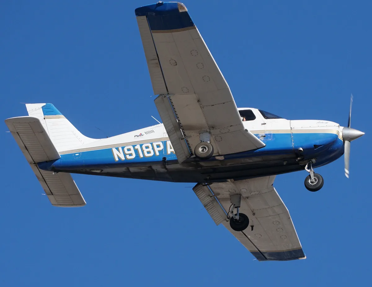 A blue and white Piper Archer III airplane is captured mid-flight against a clear blue sky, its landing gear down. The…
