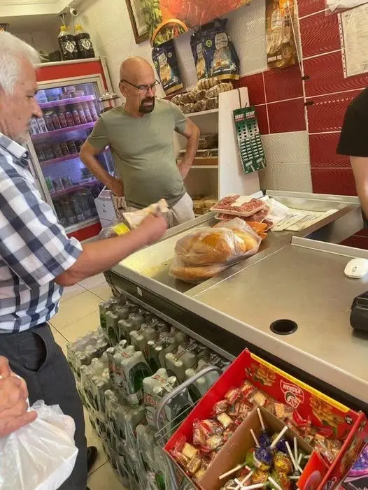 A man in a plaid shirt stands at a deli counter holding a bag, while another man in a green shirt stands behind him with…