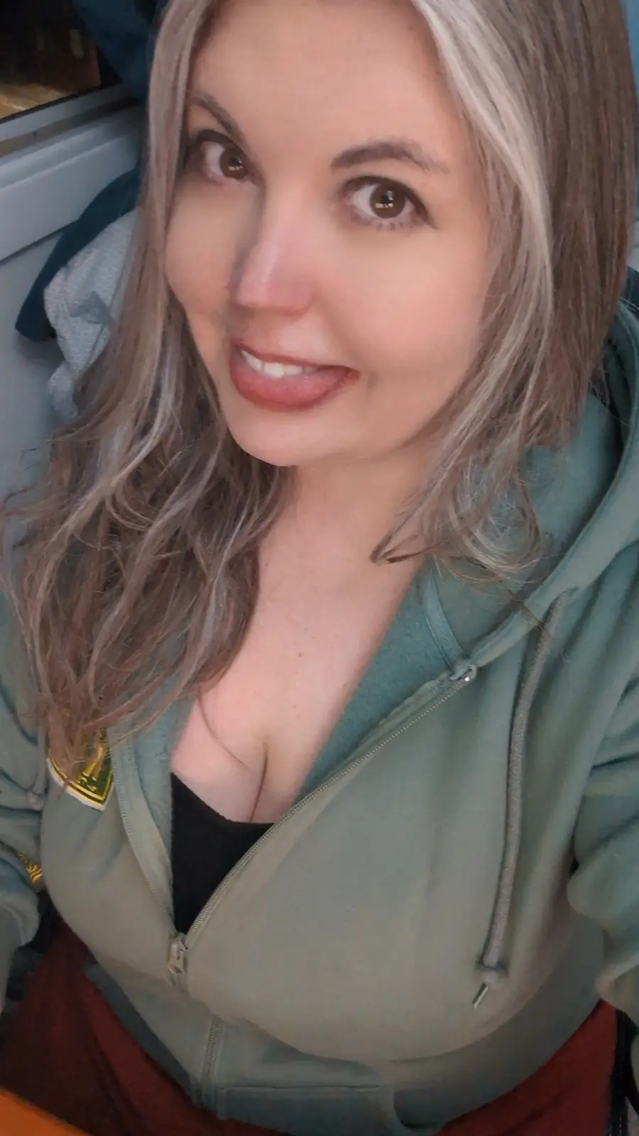 A woman with light brown and blonde hair, wearing a green zip-up hoodie over a black top, looks directly at the camera with…