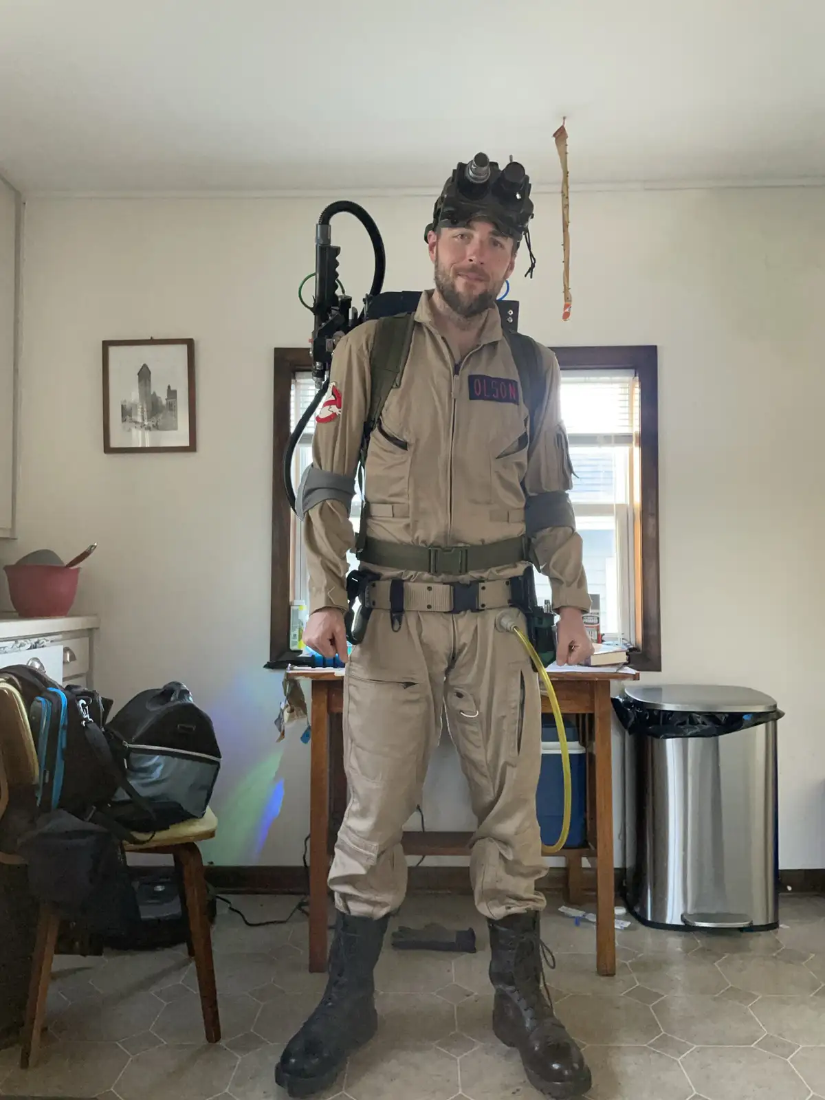 A man in a full Ghostbusters costume, complete with proton pack and helmet, stands in a messy room with hexagonal tile…