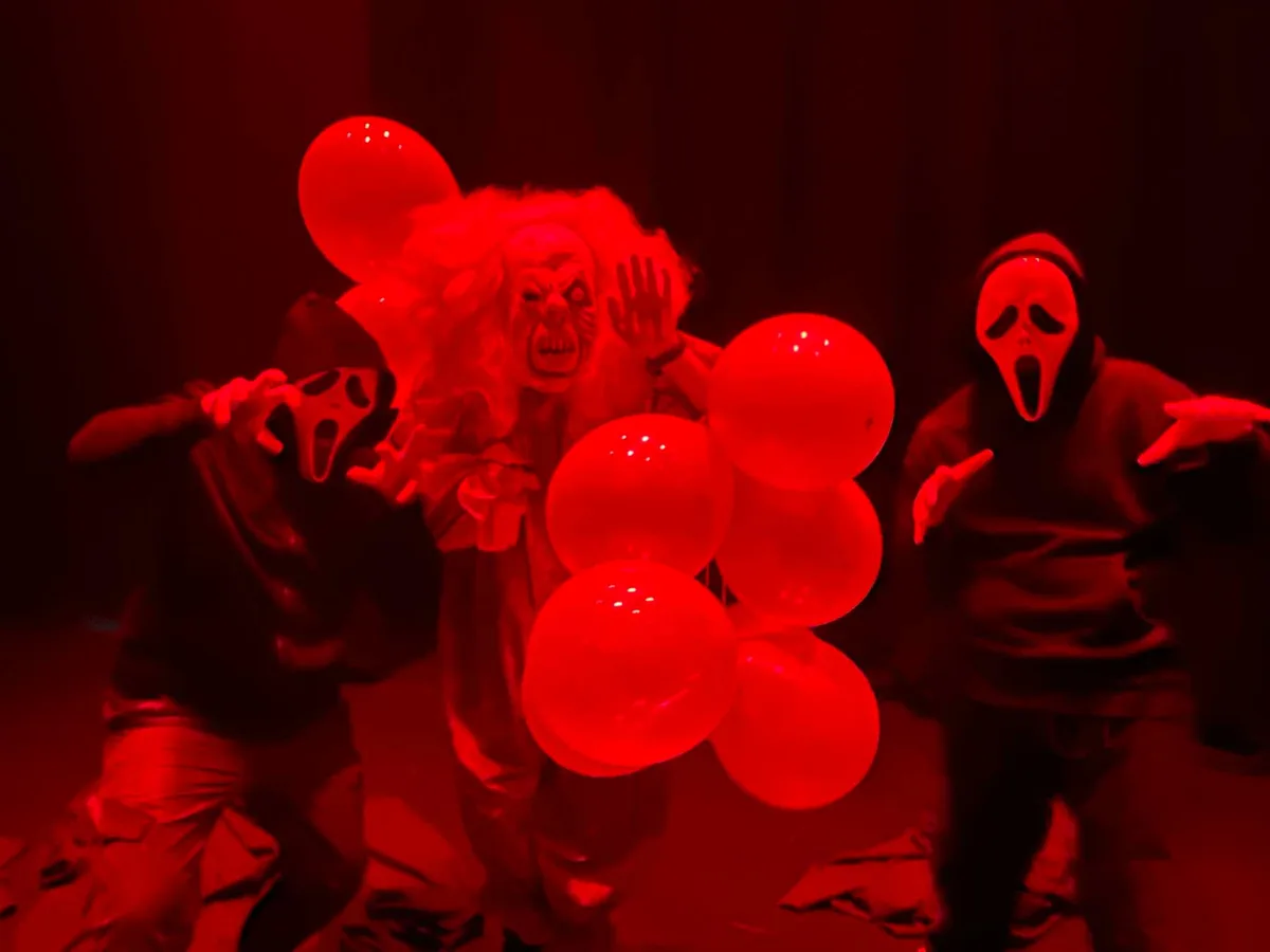 Three people in horror costumes — a clown, and two wearing Ghostface masks — are posing in a dark room bathed in red light…