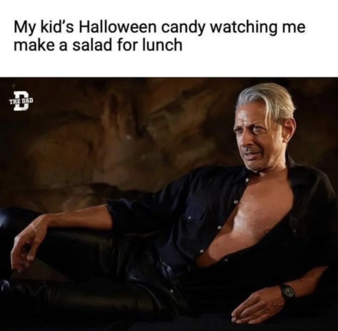 A man with gray hair, wearing an unbuttoned black shirt and black pants, sits casually on a couch with a smug, unimpressed…