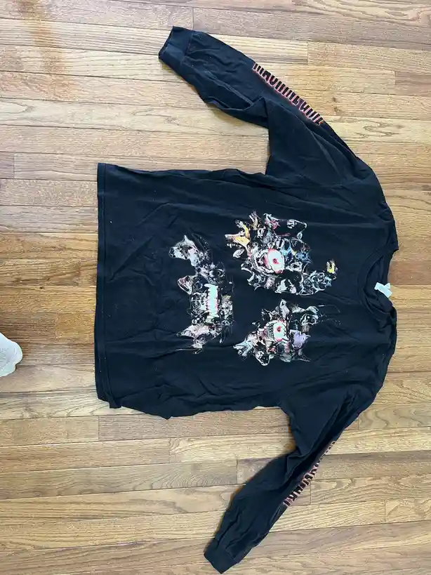 A black long-sleeved shirt lies flat on a wooden floor, featuring a large, colorful graphic of three stylized, monstrous…