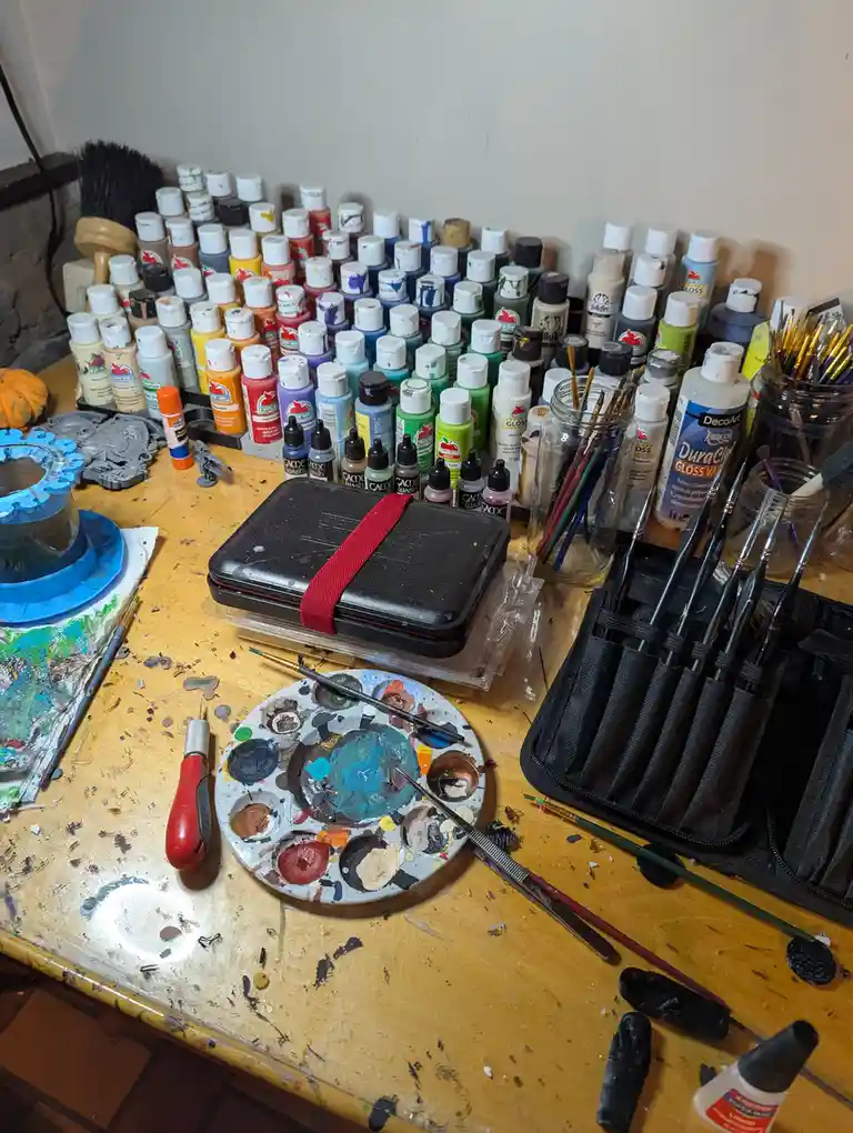 A cluttered artist's workbench is covered in paint supplies, including dozens of paint bottles, brushes in jars and a case…