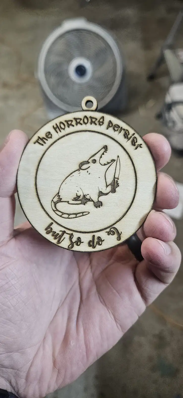A hand holds a circular wooden pendant with a laser-etched design of a cartoonish, screaming opossum holding a knife. The…