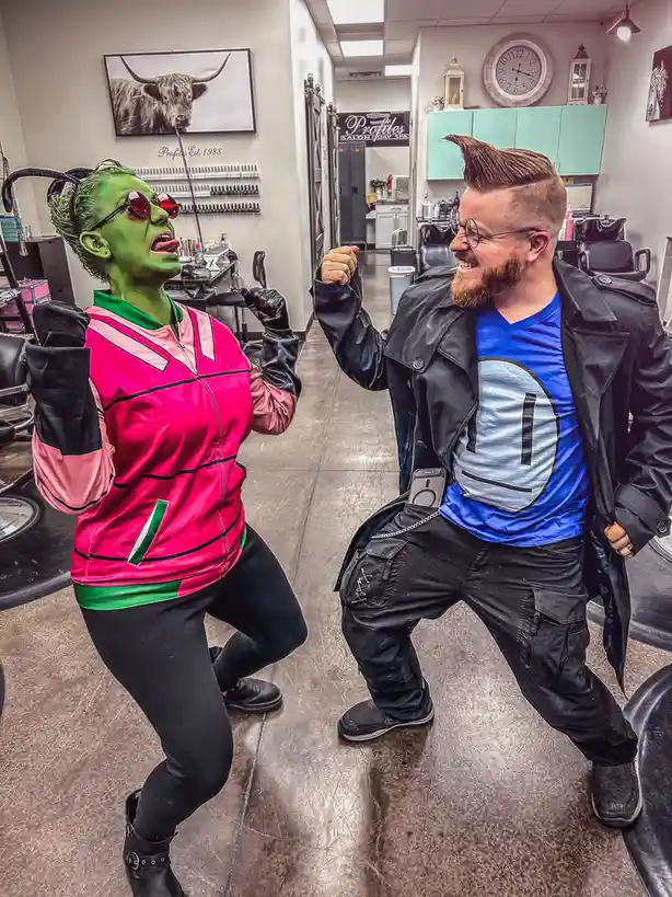 Two people in a salon pose dramatically: one is painted green with antennae and a pink jacket, the other wears a blue shirt…