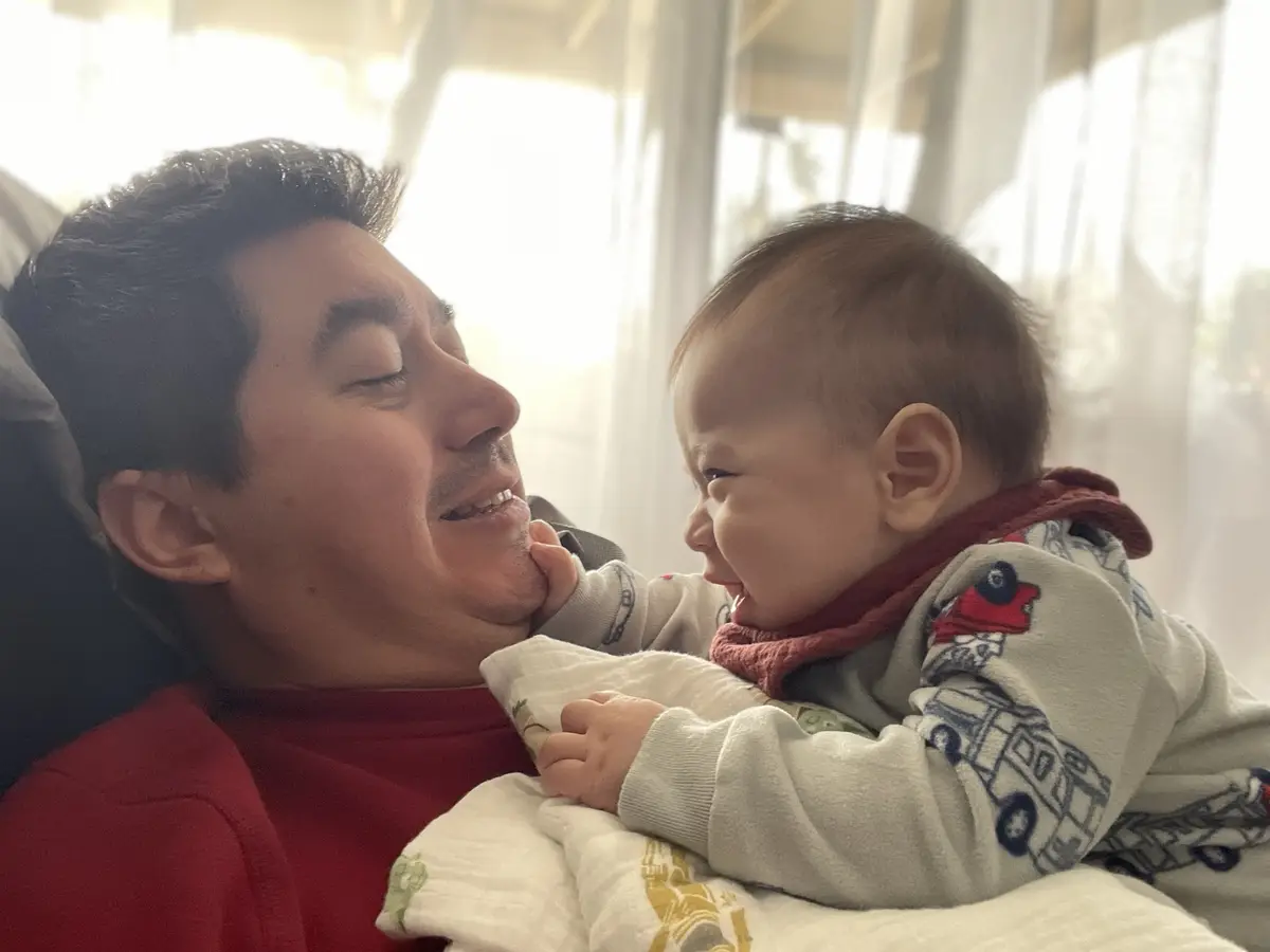 A man in a red shirt is lying down and smiling with his eyes closed while holding a baby who is laughing and looking at him…