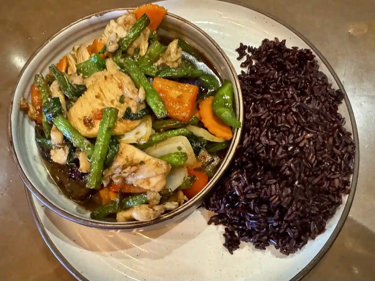 A plate holds a bowl of stir-fried chicken and vegetables next to a pile of dark purple/black rice, with overlaid text…