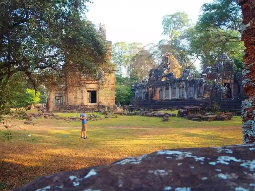 A person stands alone in a grassy field surrounded by ancient, overgrown stone ruins under bright sunlight. The ruins…