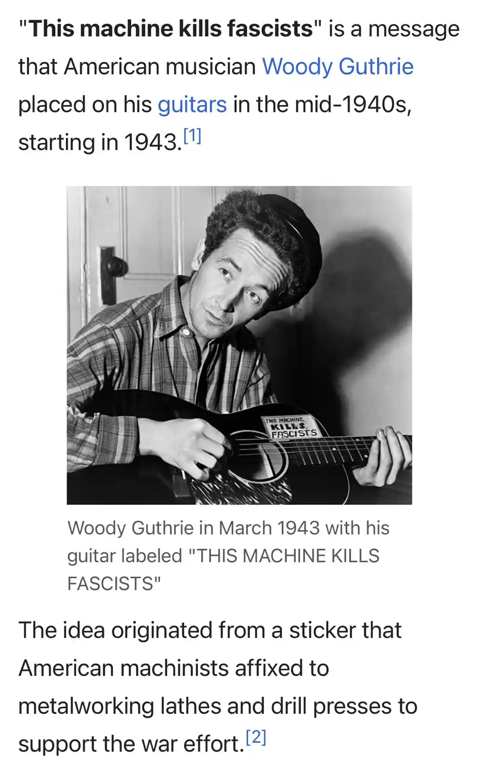 A black-and-white photo shows American musician Woody Guthrie in 1943 holding an acoustic guitar with a sticker that reads…