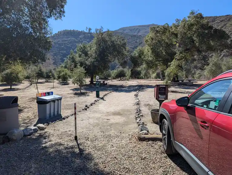 A red car is parked on a gravel path in a sunny, tree-lined park with mountains in the background. The path is lined with…