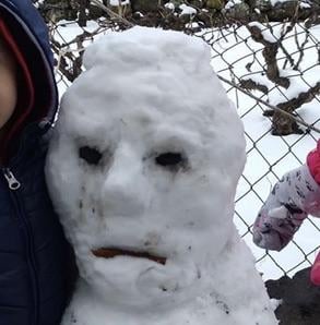 A snowman with a crudely sculpted human-like face, featuring two dark eye holes and a mouth made from a stick, stands beside…