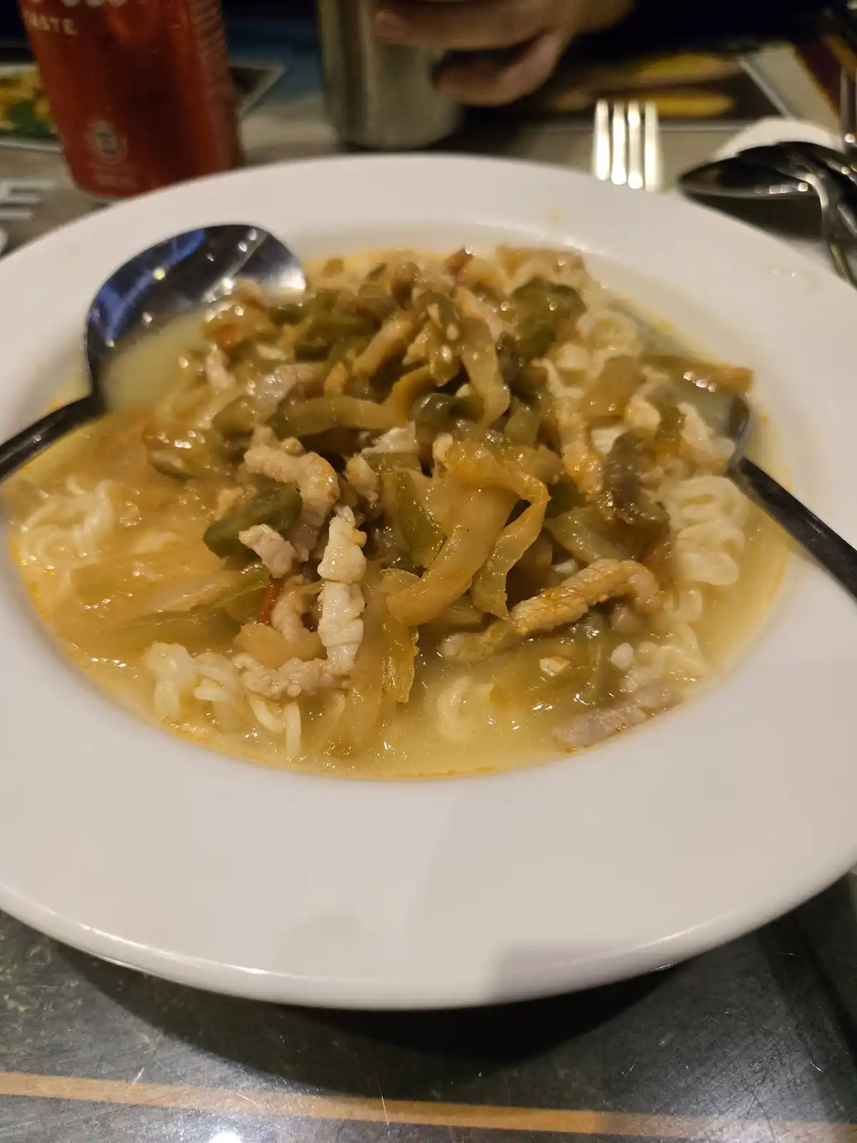 A close-up photo of a white bowl filled with noodle soup containing visible noodles, shredded meat, and green vegetables…