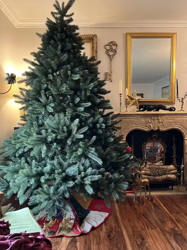 A large, unadorned artificial Christmas tree stands in a lavishly decorated living room with ornate fireplace, gold-framed…