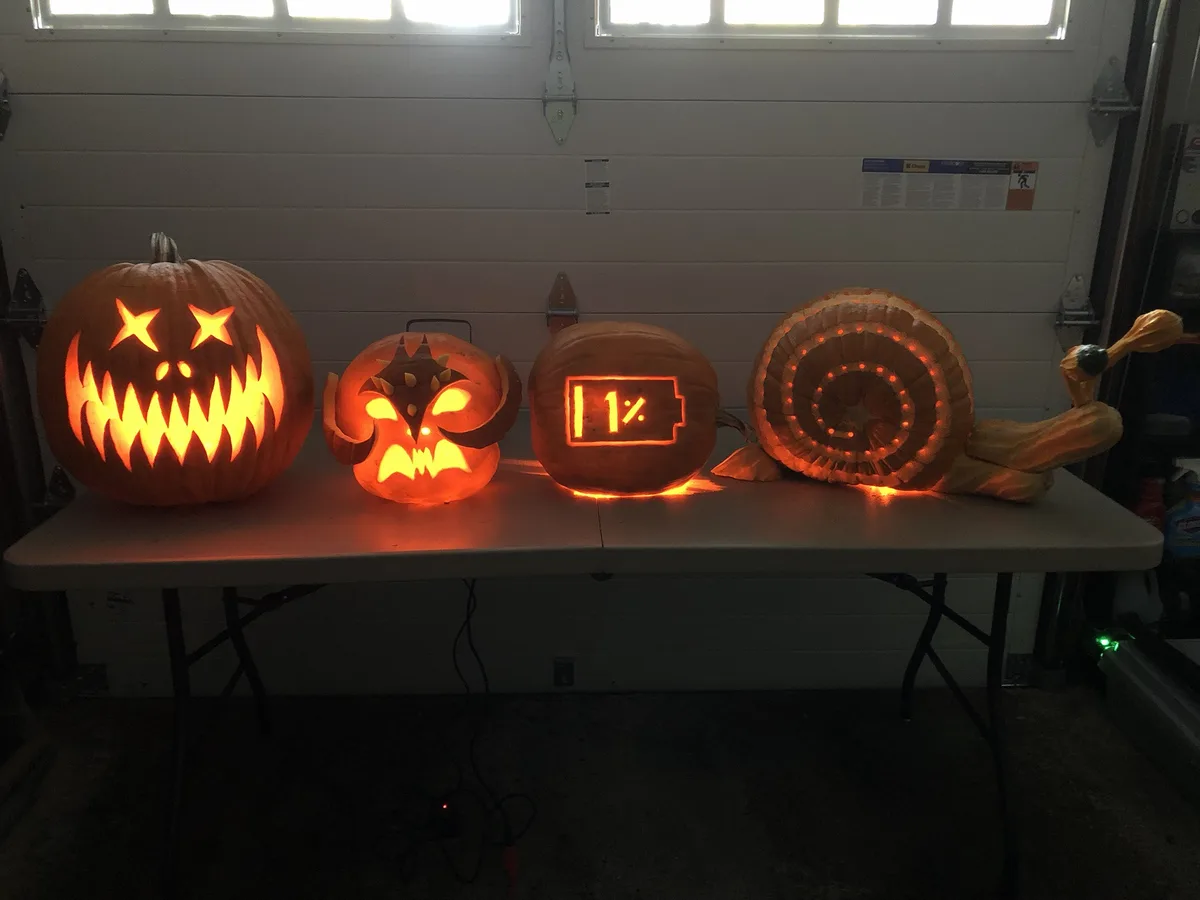 Four illuminated jack-o'-lanterns sit on a table in a garage. From left to right: a classic scary face, a demonic face, a…