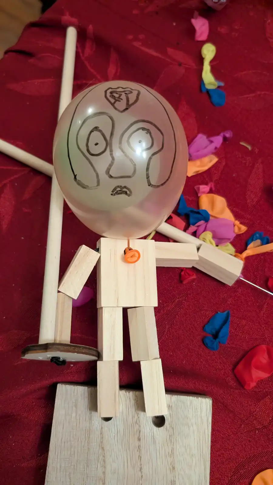 A handmade figure with a translucent balloon head, drawn with a worried face and a crown, attached to a wooden body made of…